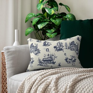 Western toile lumbar pillow in cream and navy blue placed on a light sofa with a green cushion and leafy plant in the background
