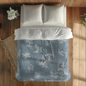 Op de afbeelding: Een blauw dekbedovertrek met een wit toile-patroon met figuren, bomen en zwanen. Het bed is opgemaakt met witte kussens en een wit laken. Het bed staat op een houten vloer.