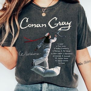 May include: Dark gray t-shirt featuring the text "Conan Gray" in white script above an illustration of a person in a sailor outfit. The shirt also lists song titles from the album "Wishbone".