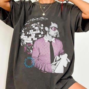 Puede incluir: Camiseta gris oscuro con un gráfico de un hombre con un suéter rosa y gafas de sol, con una bola de discoteca y texto. El texto dice "Kiss the Discasionally Time". La camiseta tiene mangas cortas y un corte holgado.