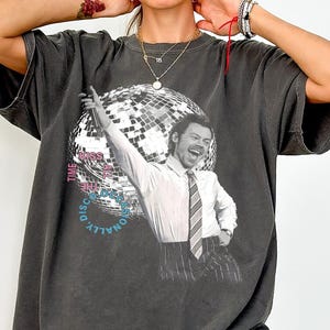 Puede incluir: Camiseta gris oscuro con una imagen en blanco y negro de un hombre con camisa blanca y corbata, con un fondo de bola de discoteca. El texto en la camiseta dice "TIME TO KISS THE DISCO PERSONALLY".