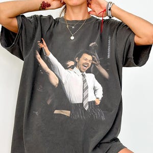 Puede incluir: Camiseta gris oscuro de gran tamaño con un gráfico de un hombre con camisa blanca, corbata a rayas y pantalón de raya diplomática, sosteniendo un micrófono y sonriendo. La imagen está impresa en la parte delantera de la camiseta.