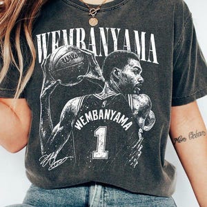 May include: Dark gray t-shirt featuring a black and white graphic of a basketball player holding a basketball. The text "WEMBANYAMA" is above the player, and the player's jersey number "1" is visible.