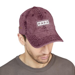 Puede incluir: Gorra de béisbol burdeos con la palabra "PAPA" en un rectángulo blanco. El texto "EST. 1973" está debajo. La gorra tiene una visera curva y un aspecto vintage desgastado.