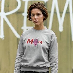 Puede incluir: Una sudadera gris claro con la palabra "Mom" en escritura roja y un gráfico de corazón rosa con una corona. La sudadera de manga larga tiene cuello redondo. El fondo es una pared con graffiti blanco.