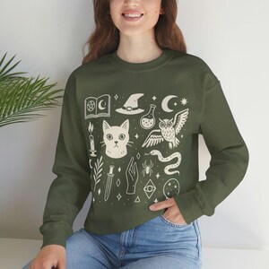 Puede incluir: Sudadera verde con un gráfico color crema que presenta un gato, un búho, una serpiente, un sombrero de bruja, una botella de poción y otros símbolos místicos. La parte superior de manga larga es de estilo casual y cómodo.