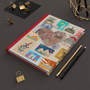 Puede incluir: Un diario con encuadernación roja y una cubierta adornada con sellos postales antiguos, incluidos sellos de París y Londres. Un recorte en forma de corazón revela un diseño de mapa. Lápices dorados y negros y clips dorados están cerca.