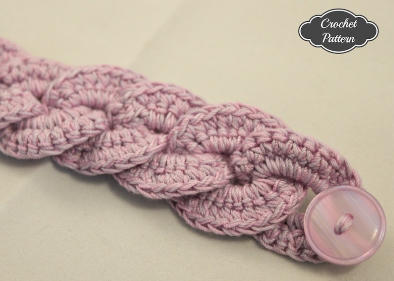 K&ouml;nnte beinhalten: Ein rosa geh&auml;keltes Stirnband mit einem rosa Knopf. Das Stirnband ist mit einem einfachen Stichmuster gearbeitet und hat einen geflochtenen Look.