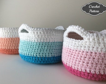 May include: Three crocheted baskets with handles. The baskets are white with a band of color at the bottom. The colors are peach, teal and turquoise, and pink and red.
