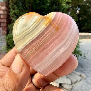 Puede incluir: Una piedra en forma de corazón con capas de tonos rosa, crema y amarillo. La superficie pulida revela un patrón natural a rayas. La piedra se sostiene en una mano, mostrando su tamaño y forma. Este artículo decorativo es un regalo único.