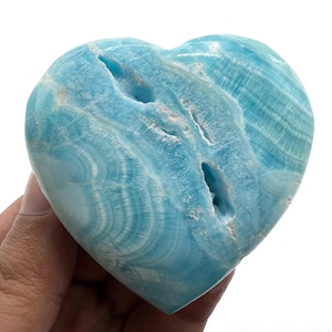 Könnte beinhalten: Ein herzförmiger, hellblauer Stein mit strukturierter Oberfläche und natürlichen Löchern. Der Stein hat ein geschichtetes Aussehen mit weißen und blauen Bändern. Das Herz wird in einer Hand vor weißem Hintergrund gehalten.