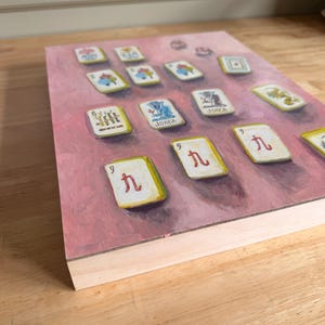 May include: A rectangular painting of Mahjong tiles on a pink background. The tiles are white with red and black symbols and characters. The painting has a wooden frame and is displayed at an angle, showing the side profile of the frame.