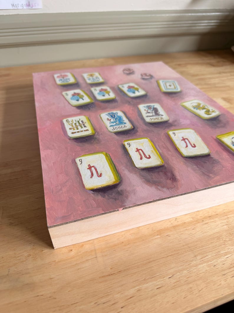 May include: A rectangular painting of Mahjong tiles on a pink background. The tiles are white with red and black symbols and characters. The painting has a wooden frame and is displayed at an angle, showing the side profile of the frame.