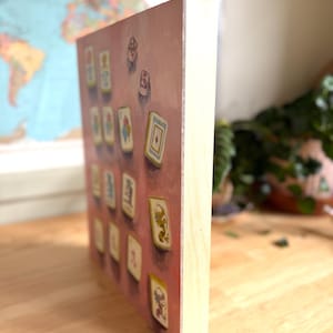 May include: A wooden block print featuring a collection of illustrated playing cards against a pink background. The cards have various designs and colors, with a light wood frame. The artwork is displayed on a wooden surface.