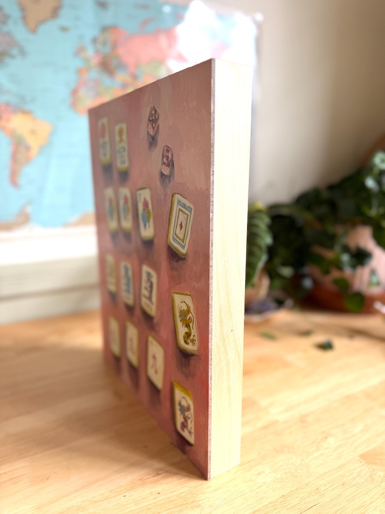 May include: A wooden block print featuring a collection of illustrated playing cards against a pink background. The cards have various designs and colors, with a light wood frame. The artwork is displayed on a wooden surface.