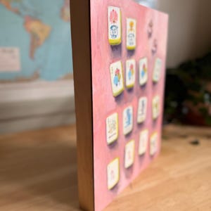May include: A pink canvas print featuring a collection of illustrated cards. The cards have a white border and are arranged on a pink background. The wooden frame adds a natural contrast to the artwork.