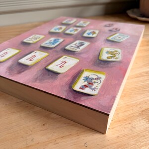 May include: A painting of Mahjong tiles on a pink background. The tiles are white with various colorful illustrations and Chinese characters. The wooden frame is visible, and the artwork is displayed at an angle.