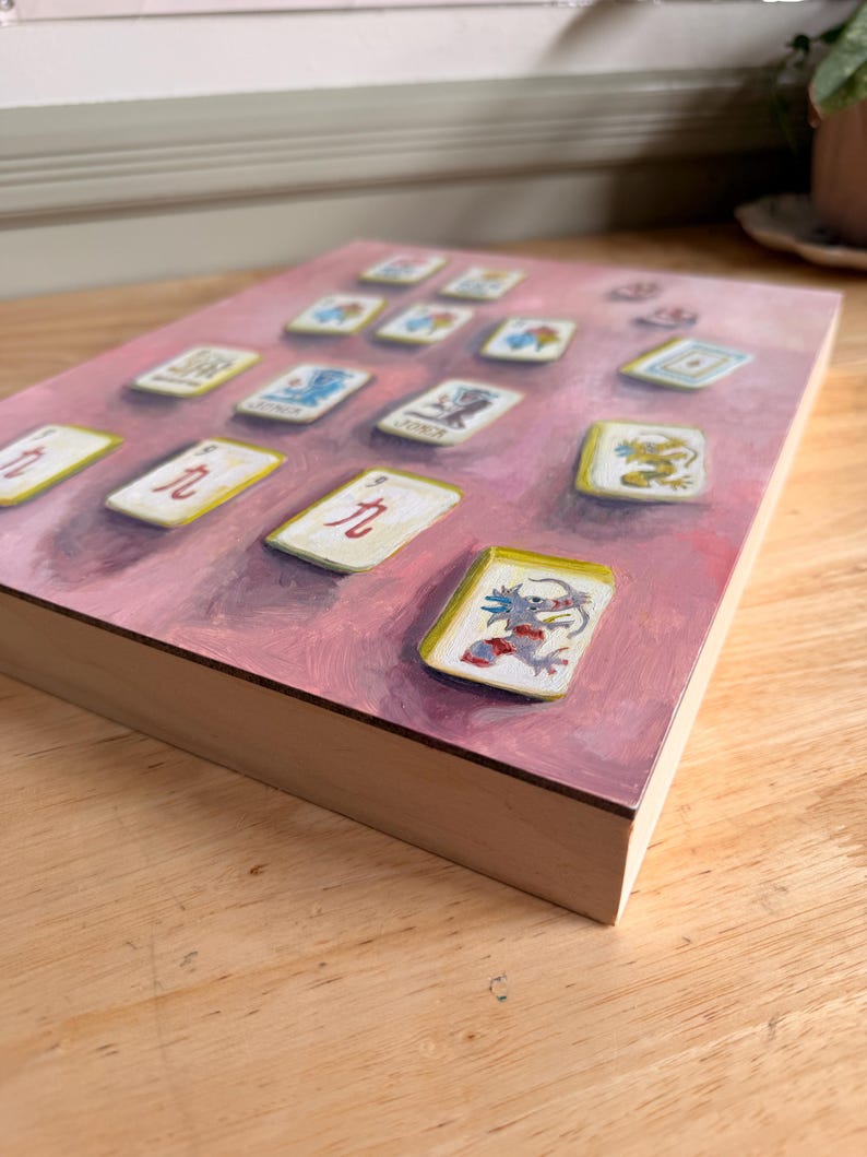 May include: A painting of Mahjong tiles on a pink background. The tiles are white with various colorful illustrations and Chinese characters. The wooden frame is visible, and the artwork is displayed at an angle.