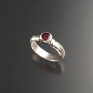 Puede incluir: Un anillo de plata con una piedra preciosa roja engastada en el centro. La banda está decorada con un patrón floral.