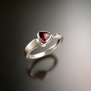 May include: A silver ring with a triangular, faceted, deep red gemstone. The gemstone is set in a bezel setting, and the band has a simple, modern design. The ring is displayed on a reflective surface.
