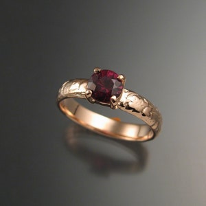 Puede incluir: Un anillo de oro rosa con una piedra preciosa roja oscura engastada en una banda texturizada.