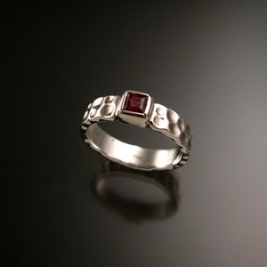 Puede incluir: Un anillo de plata con textura martillada y una gema roja oscura cuadrada. La banda del anillo tiene un patrón texturizado, y la gema está engastada en un bisel cuadrado. El anillo se muestra sobre un fondo oscuro.