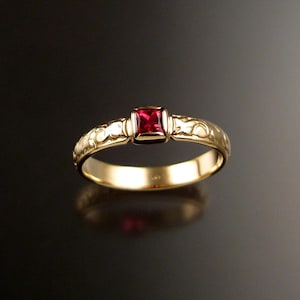 Puede incluir: Un anillo de banda de oro con una piedra preciosa roja cuadrada engastada en el centro. La banda está decorada con un diseño floral.