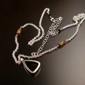 Op de afbeelding: Een zilveren ketting met een driehoekige hanger met een zwarte steen. De ketting is versierd met kleine amberkleurige en witte kralen.