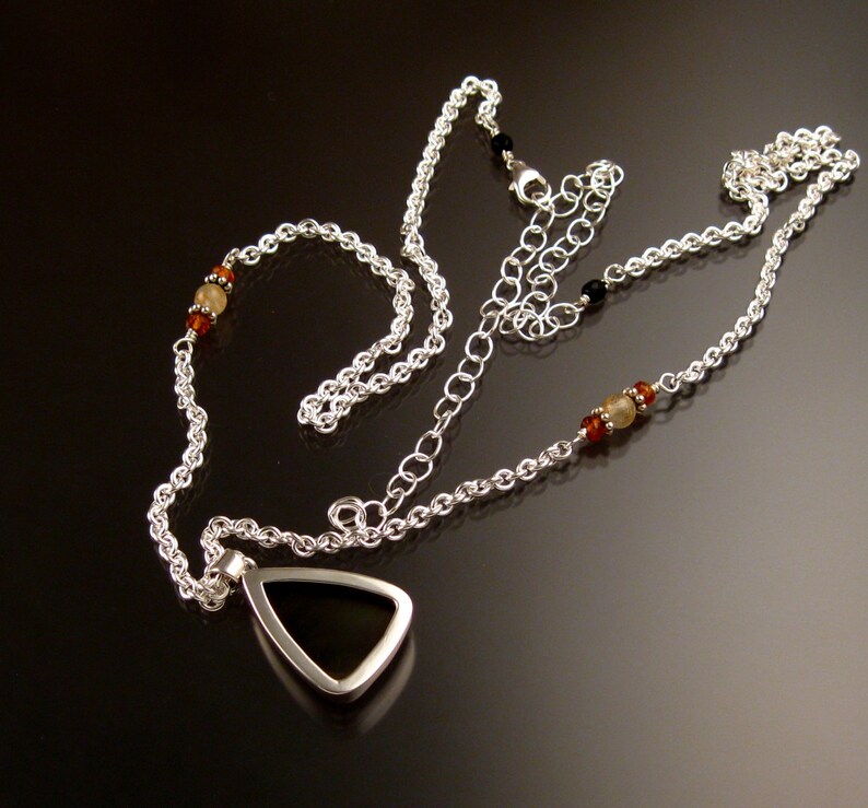 Op de afbeelding: Een zilveren ketting met een driehoekige hanger met een zwarte steen. De ketting is versierd met kleine amberkleurige en witte kralen.