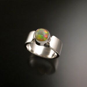 May include: A silver ring with a round, iridescent opal gemstone set in a bezel setting. The ring has a wide band with a slightly rounded edge.