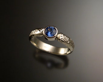 Puede incluir: Un delicado anillo de banda de oro con una piedra preciosa de zafiro azul engastada en un engaste de bisel. La banda está adornada con un intrincado diseño floral.