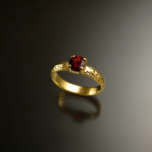Puede incluir: Un anillo de oro con una piedra preciosa roja engastada en el centro. La banda está decorada con un diseño floral.