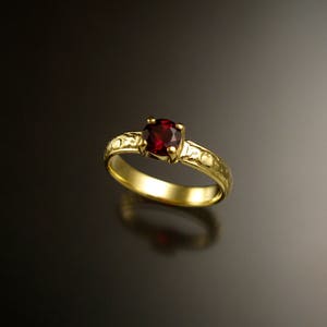 Puede incluir: Un anillo de oro con una piedra preciosa roja engastada en el centro. La banda del anillo está decorada con un patrón texturizado.