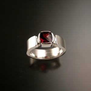 Puede incluir: Un anillo de plata con una piedra preciosa roja engastada en un bisel cuadrado. El anillo tiene una banda ancha con un borde ligeramente redondeado.