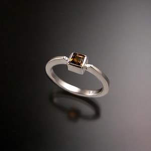 Puede incluir: Un anillo de plata con una piedra preciosa marrón de forma cuadrada engastada en el centro.