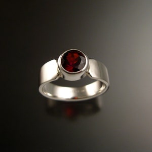 Puede incluir: Un anillo de plata con una piedra preciosa redonda roja engastada en un engaste de bisel. El anillo tiene un diseño simple y moderno.