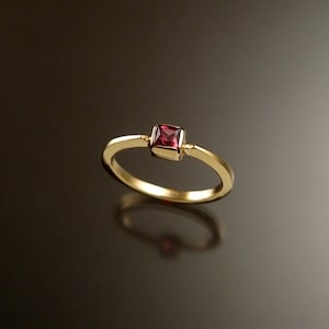 Puede incluir: Un anillo de banda de oro simple con una piedra preciosa roja de forma cuadrada.