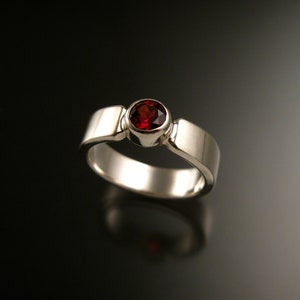 May include: A silver ring with a red gemstone set in a bezel setting. The ring has a simple, modern design.
