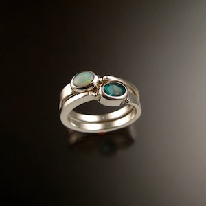 May include: A silver ring with two oval-shaped gemstones. The larger stone is a white opal, and the smaller stone is a green emerald. The ring is set on a black background.