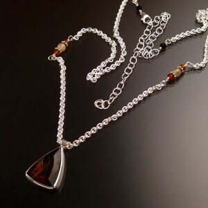 Op de afbeelding: Een zilveren ketting met een driehoekige hanger met een bruine en zwarte steen. De ketting heeft kleine amberkleurige en zwarte kralen.