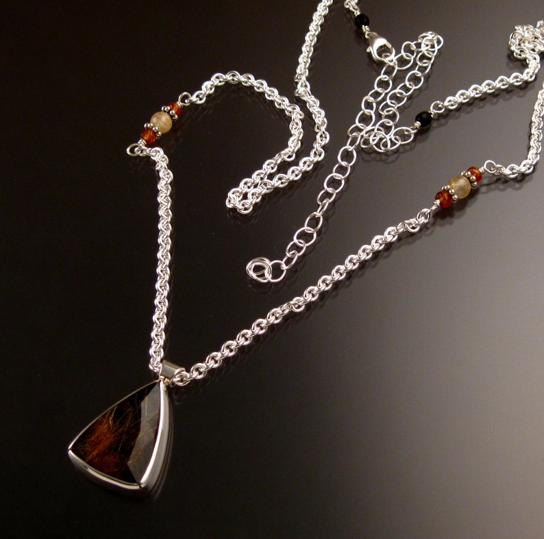 Op de afbeelding: Een zilveren ketting met een driehoekige hanger met een bruine en zwarte steen. De ketting heeft kleine amberkleurige en zwarte kralen.