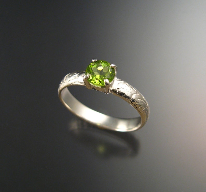 May include: A silver ring with a green gemstone set in a four-prong setting. The band is engraved with a floral design.