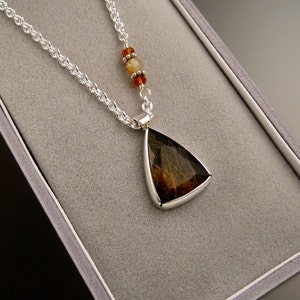 Op de afbeelding: Een zilveren ketting met een driehoekige hanger met een bruine en zwarte steen. De hanger is ingelegd in zilver en heeft een gladde, gepolijste afwerking. De ketting heeft kleine, ronde kralen in tinten oranje en bruin.