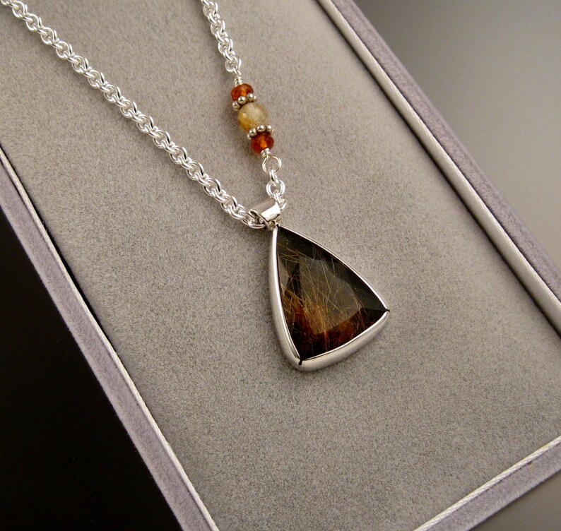 Op de afbeelding: Een zilveren ketting met een driehoekige hanger met een bruine en zwarte steen. De hanger is ingelegd in zilver en heeft een gladde, gepolijste afwerking. De ketting heeft kleine, ronde kralen in tinten oranje en bruin.