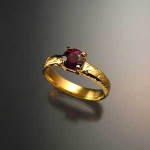 Puede incluir: Un anillo de oro con una piedra preciosa de color rojo intenso engastada en una montura de cuatro garras. La banda está texturizada con un diseño floral.