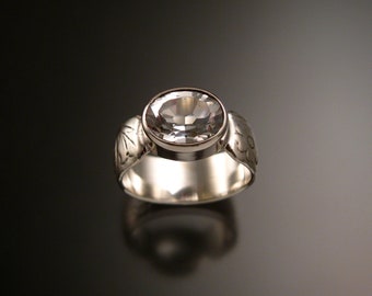 May include: A silver ring featuring a large, oval, clear gemstone. The wide band has etched floral patterns on the sides. The ring is set against a dark, reflective background.