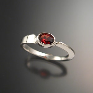 May include: A silver ring featuring a red gemstone set in a bezel setting. The ring has a simple, modern design.