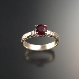 May include: A silver ring with a deep red gemstone set in the center. The band is decorated with a delicate floral pattern.
