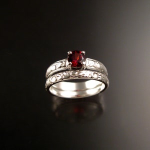 Puede incluir: Un juego de anillo de compromiso y alianza de plata. El anillo de compromiso presenta una piedra preciosa redonda de color rojo intenso. Ambos anillos tienen un diseño ornamentado de inspiración floral. Los anillos se exhiben sobre una superficie oscura y reflectante.