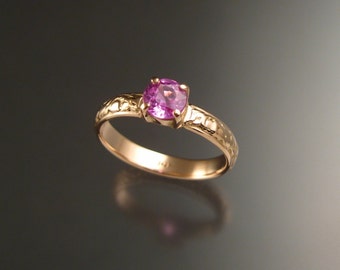Puede incluir: Un anillo de oro con una gema rosa redonda. La banda tiene un diseño detallado y texturizado. El anillo se encuentra sobre un fondo oscuro, resaltando el color de la gema y los intrincados detalles del anillo.